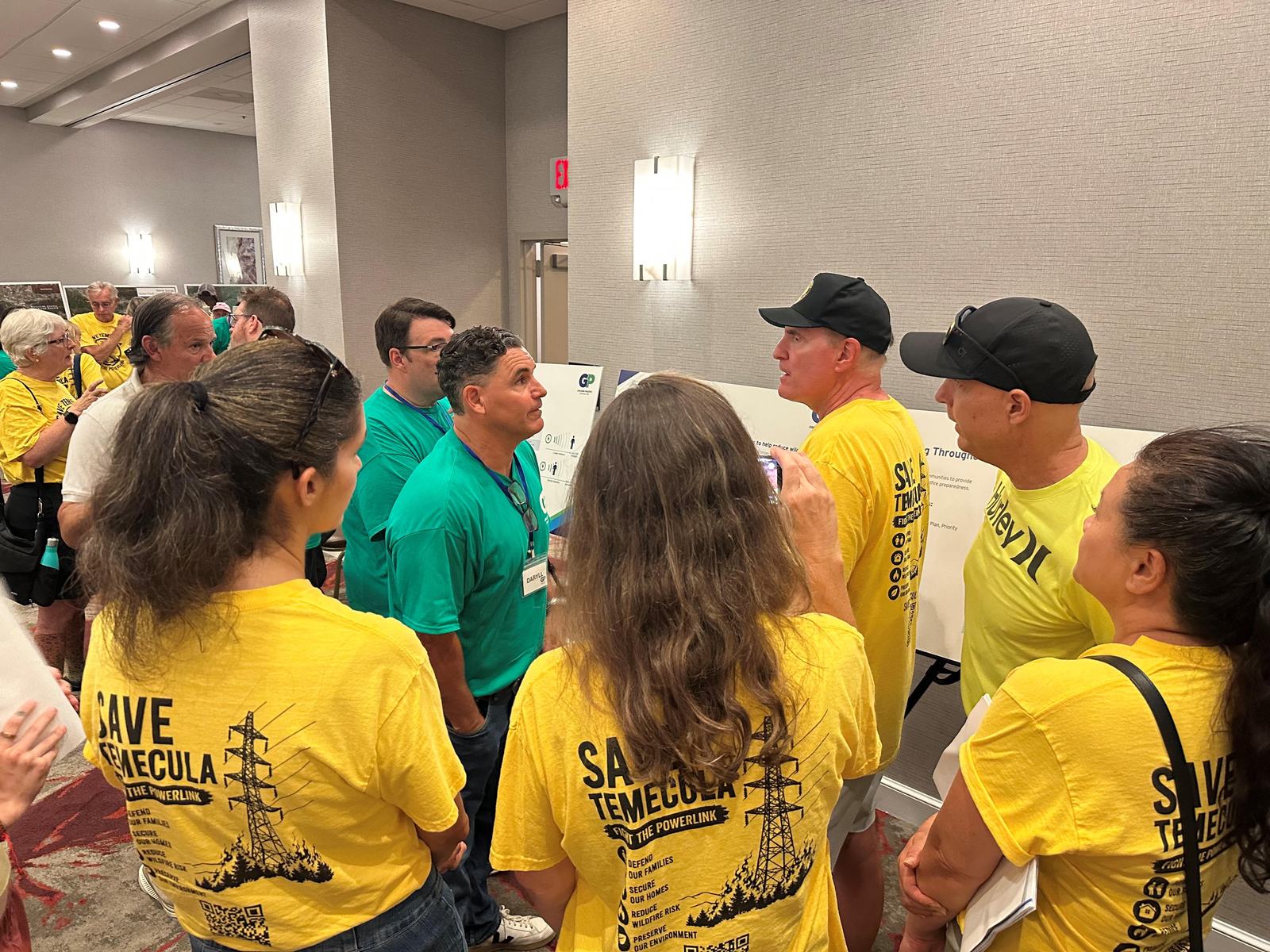 Residents in yellow "Save Temecula — Stop the Powerlink" shirts crowd shoulder to shoulder around SDG&E staff and a project poster board at the August 26 open house in Temecula.