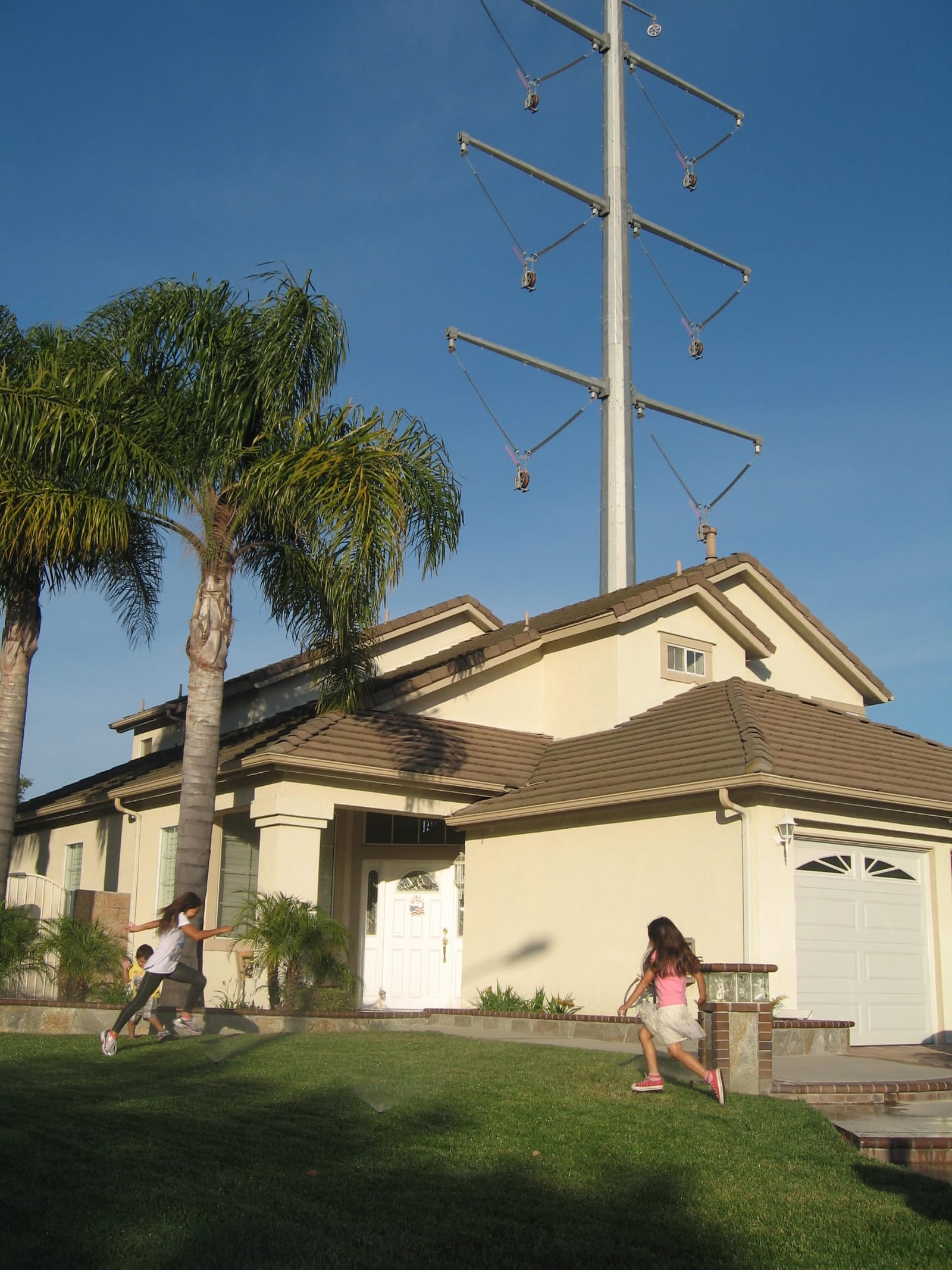 A 500 kV transmission pole standing directly behind a single-family home, with two children playing on the front lawn for scale.